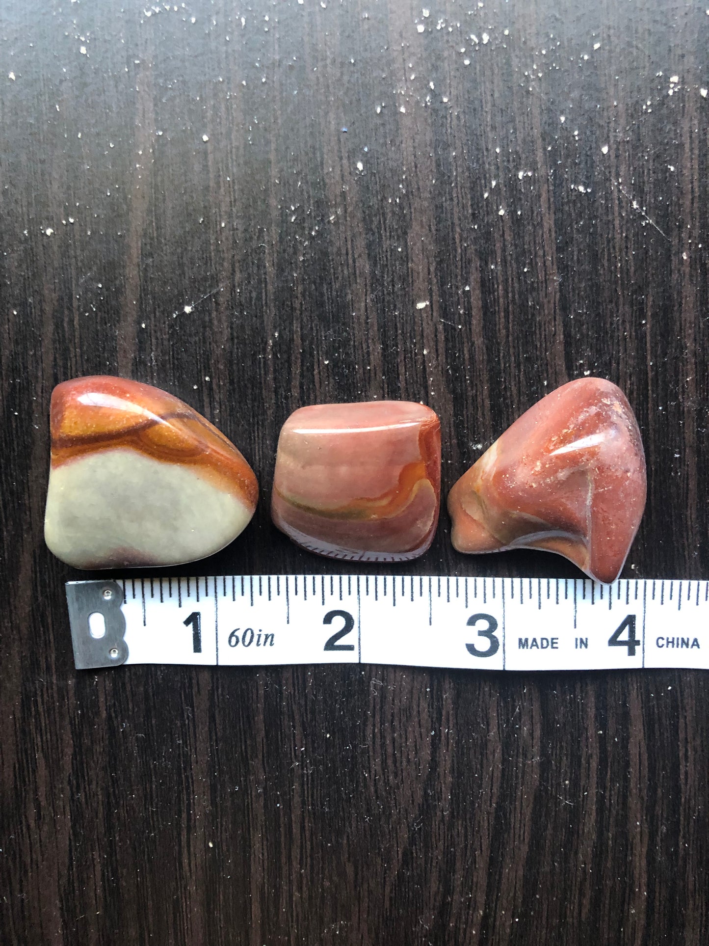 Three brown and red stones sit against a dark wood background. They are next to a white ruler showing each stone is approximately one inch wide.