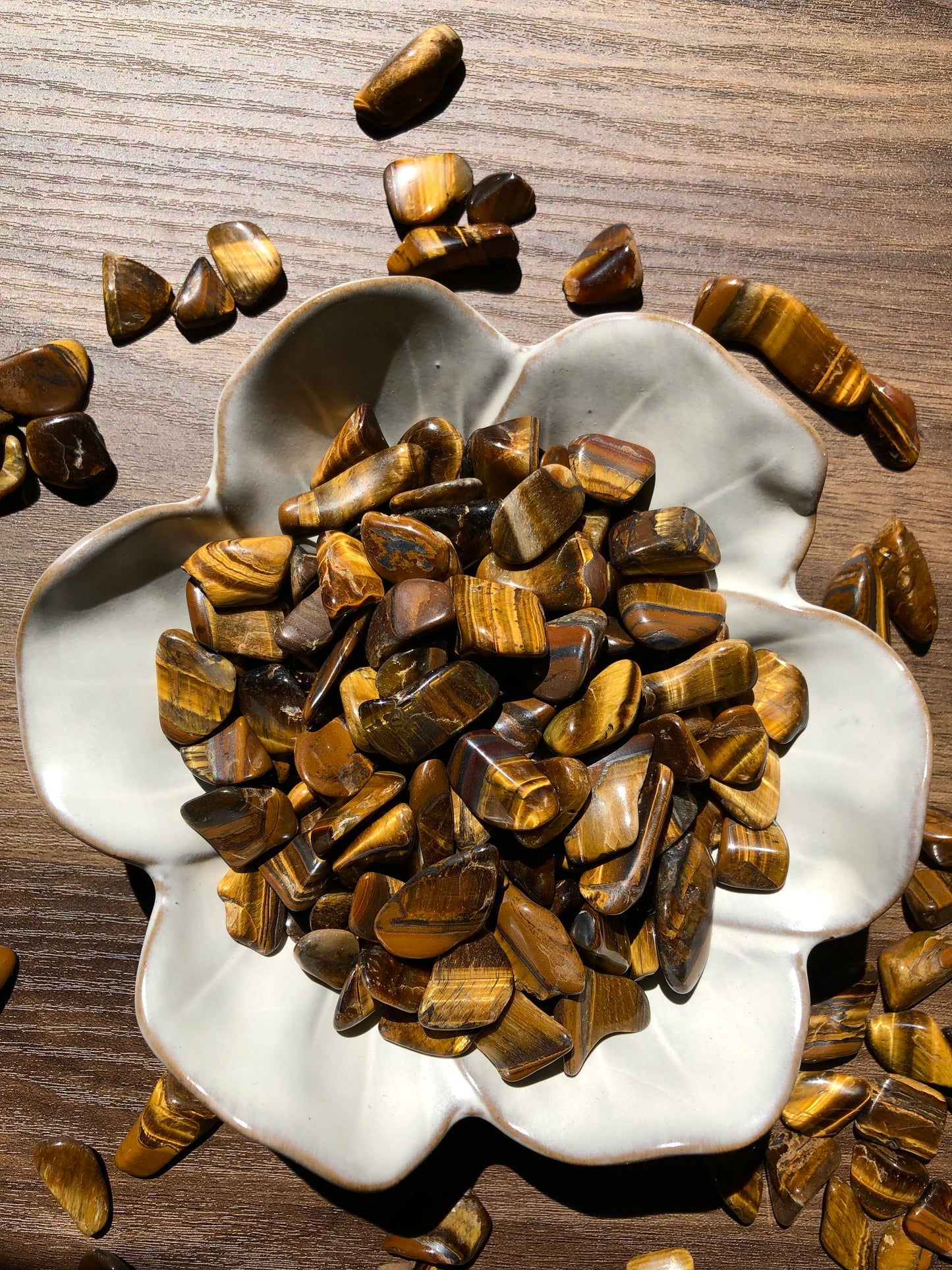 A downward shot of gold tiger eye stones sitting in a white, flower shaped bowl. There are scattered tiger eye stones around it. It sits on a dark wooden background.