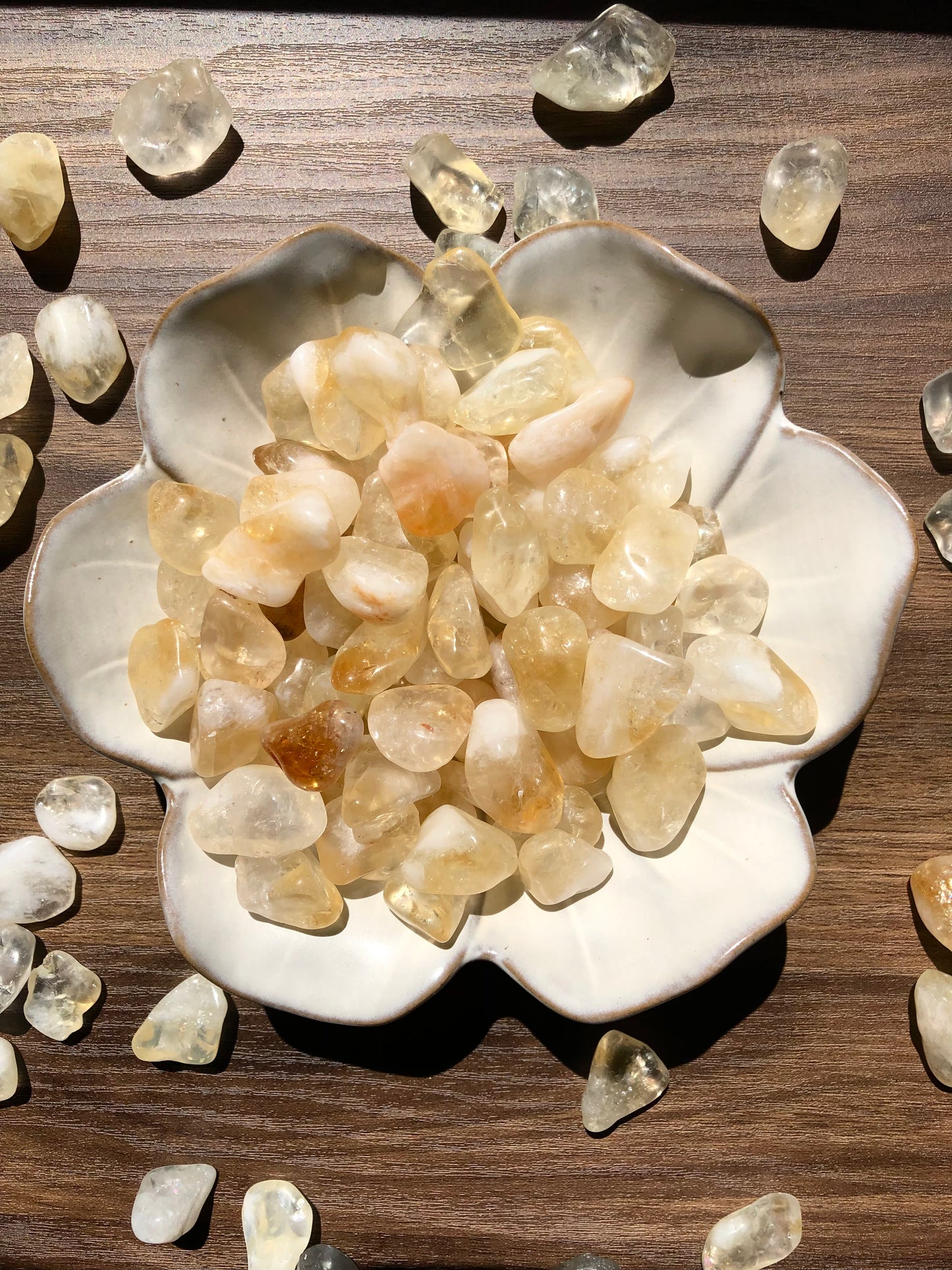 A downward picture of citrine stones sitting in a white, flower shaped bowl. There are scattered citrine stones around it. It rests on a dark wooden background.
