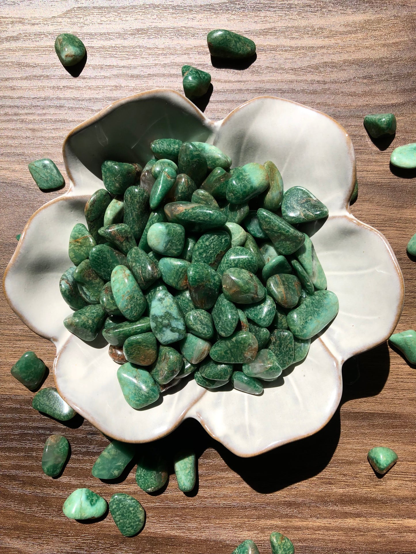 A downward picture of green quartz stones in a white, flower shaped bowl. There are scattered green quartz around it. It sits on a dark wooden background.