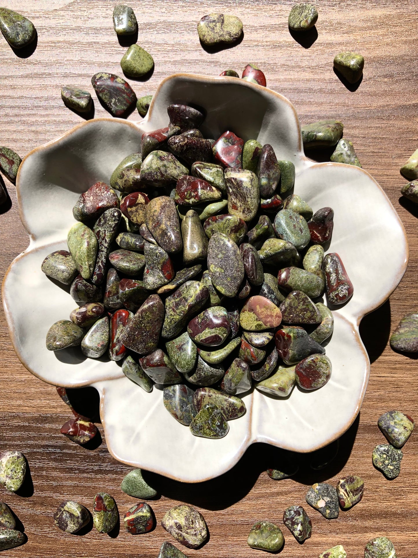 A downward picture of dragon's blood stones that sits in a white, flower shaped bowl. There are scattered dragon's blood stones around it. It sits on a dark wooden background.