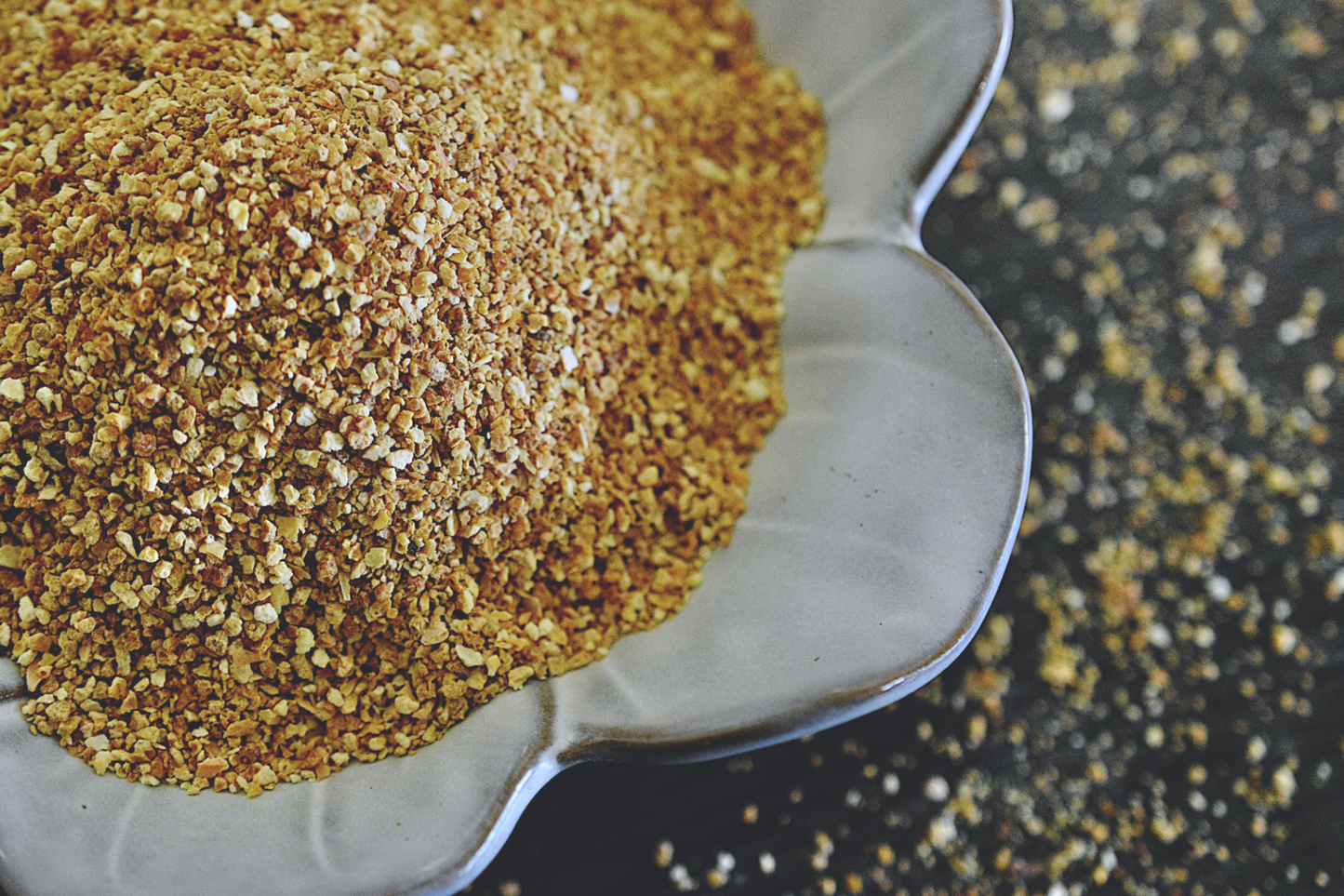 A close up view of an assortment of orange peel granules in a white, flower shaped dish on a wooden surface. The granules are a soft orange and yellow color. Small granules are scattered about in the background.