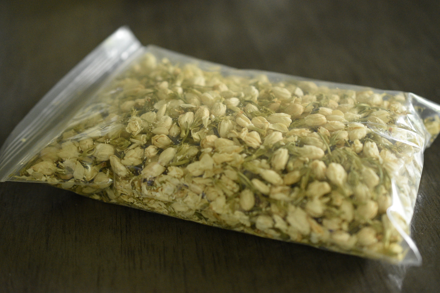 A plastic bag filled with jasmine buds sits on a wooden surface to show the packaged product.. The jasmine buds are a soft, creamy white.