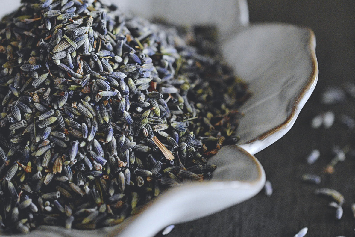 A close up view of an assortment of lavender petals rests in a white, flower shaped dish on a wooden surface. The lavender is a soft purple with hints of blue. There are lavender petals scattered about in the background.