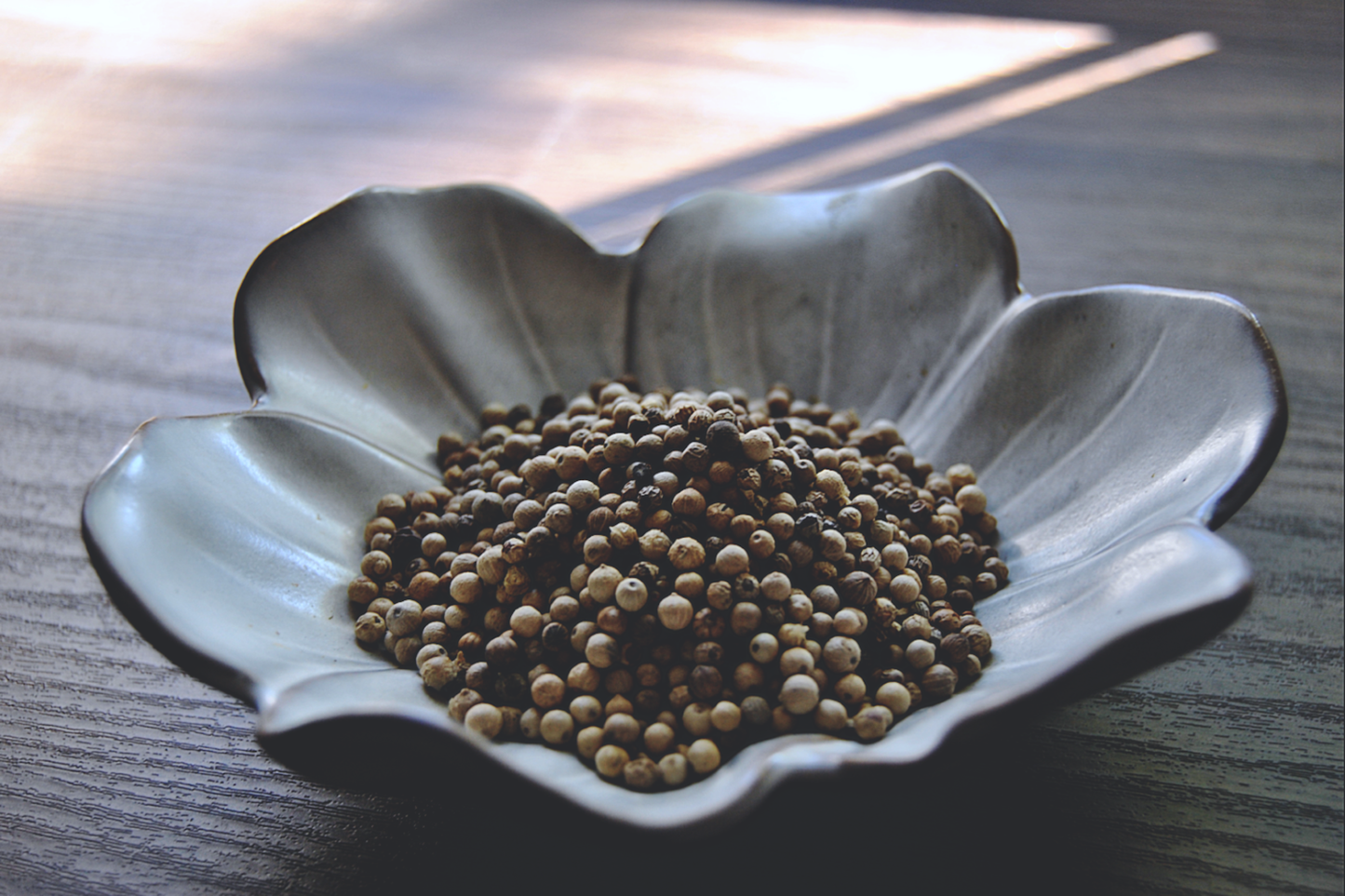 An assortment of white peppercorns rests in a white, flower shaped dish on a wooden surface. Some of the white peppercorns are slightly darker than white.