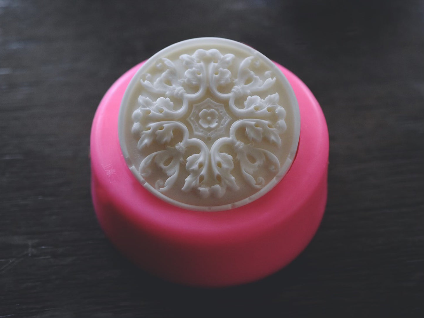 A round bar of soap with an elegant, filigree design rests in a round soap mold on a wooden surface. The mold is pink.