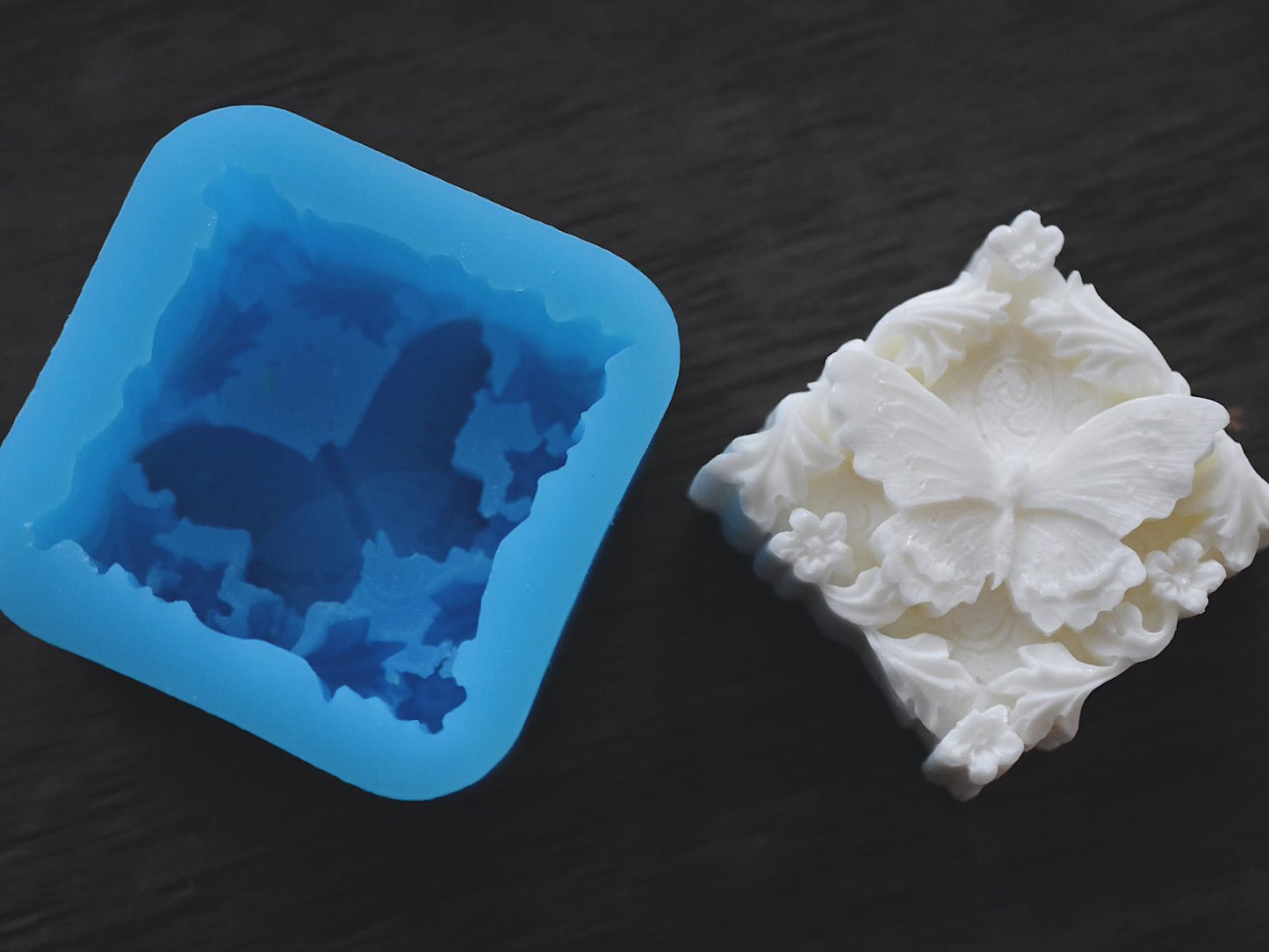 A square bar of soap with a butterfly and flower design sits next to the butterfly mold on a wooden surface. The bar of soap is white and the mold is blue.