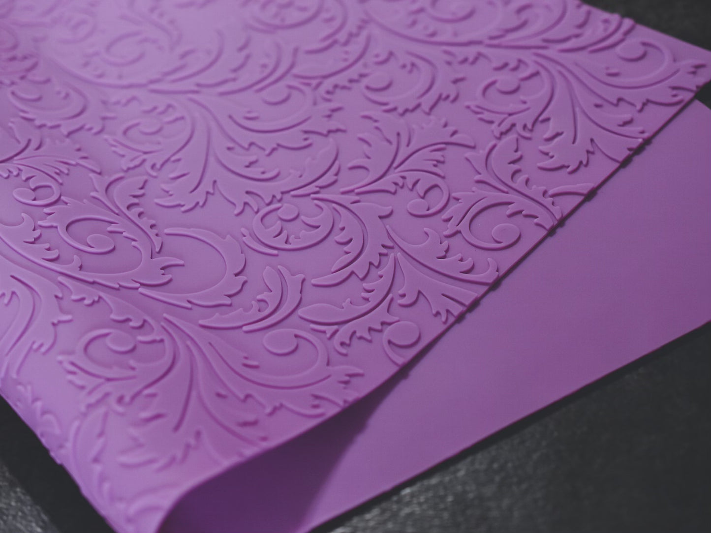A textured purple mat with a floral filigree design rests on a dark surface. The mat is slightly folded on itself.