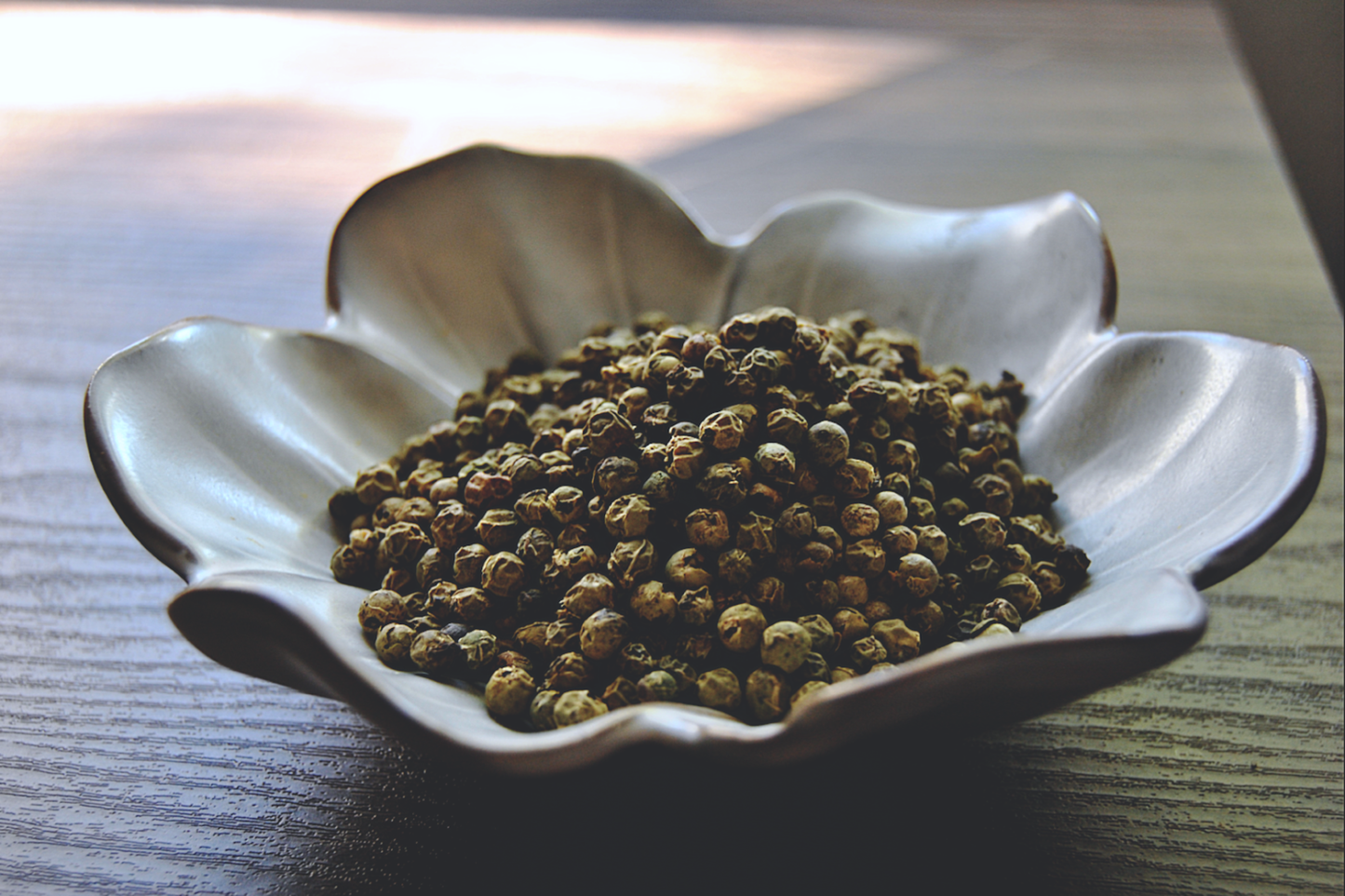 An assortment of green peppercorns rests in a white, flower shaped dish on a wooden surface.