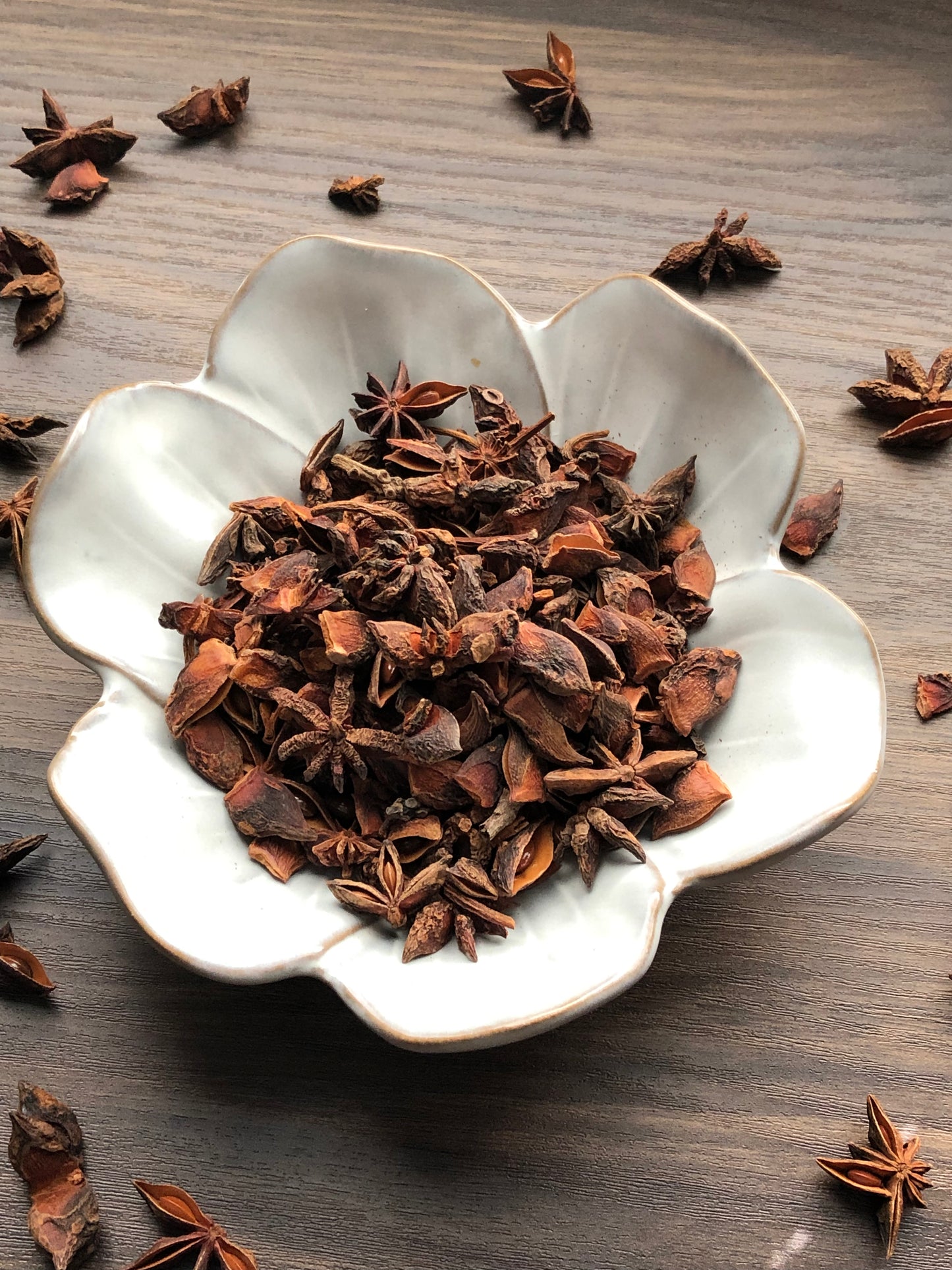 A top down view of an assortment of dried star anise pieces rests in a white, flower shaped bowl on a wooden surface. The dried flowers are dark brown in color. Most are chipped and have petals that are missing. Some flowers are scattered about in the background.