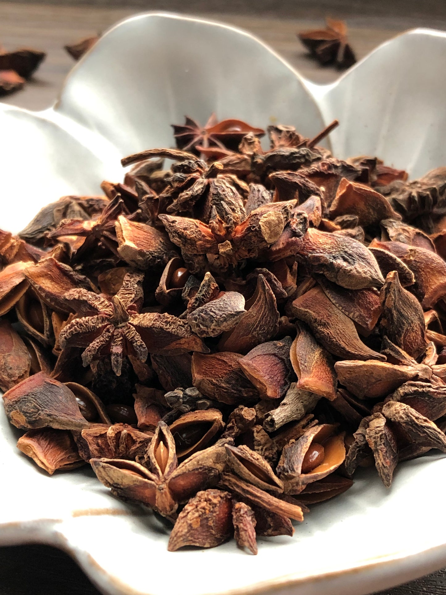A close up view of an assortment of dried star anise pieces rests in a white, flower shaped bowl on a wooden surface. The dried flowers are dark brown in color. Most are chipped and have petals that are missing. Some flowers are scattered about in the background.