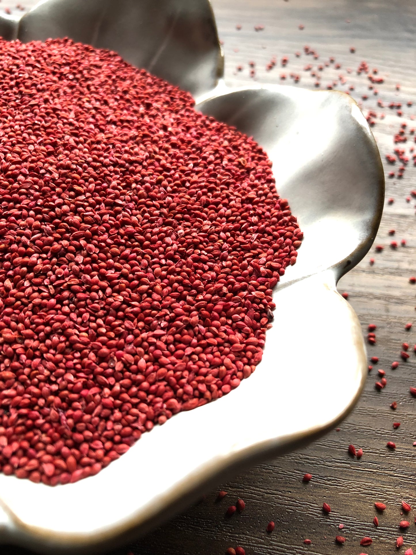 A close up view of the small and vibrant red cranberry seeds sits in a white, flower shaped dish on a wooden surface. Small seeds are scattered about in the background.