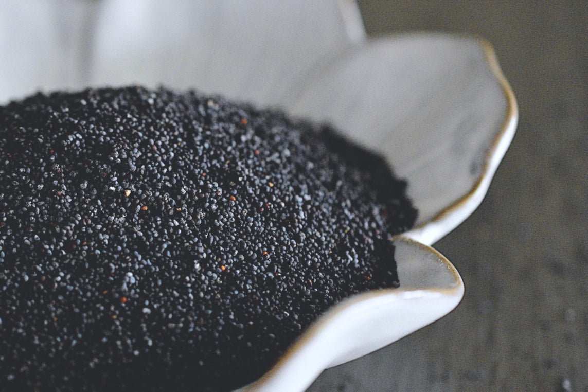 A close up view of an assortment of poppy seeds in a white, flower shaped dish on a wooden surface. The poppy seeds are dark purple, almost black in color.
