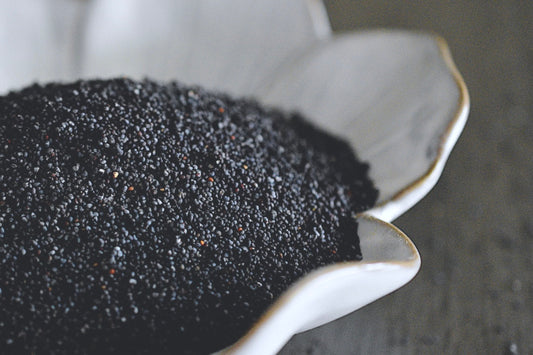 A close up view of an assortment of poppy seeds in a white, flower shaped dish on a wooden surface. The poppy seeds are dark purple, almost black in color.