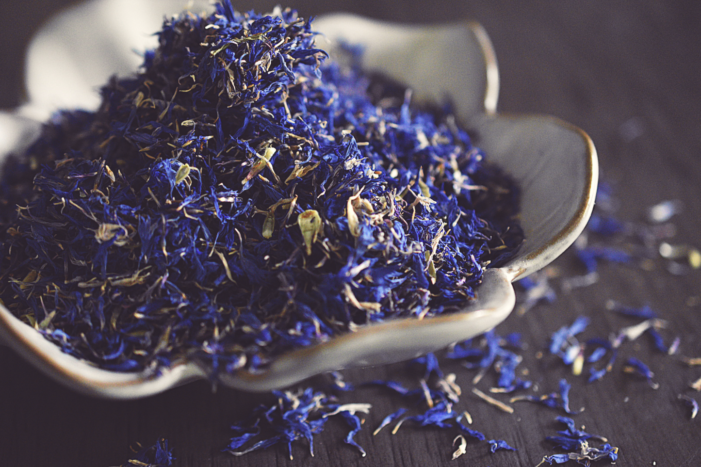 A close up photo of sprinkled blue cornflowers in a small, white, flower shaped dish on a wooden surface.