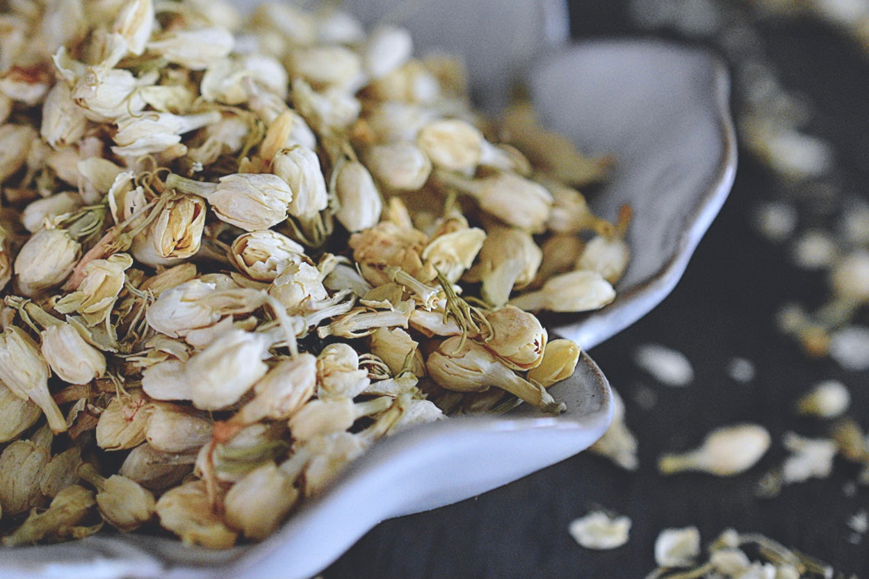 A close up view of an assortment of jasmine buds rests in a white, flower shaped dish on a wooden surface. The jasmine buds are a soft, creamy white. There are jasmine buds scattered about in the background.