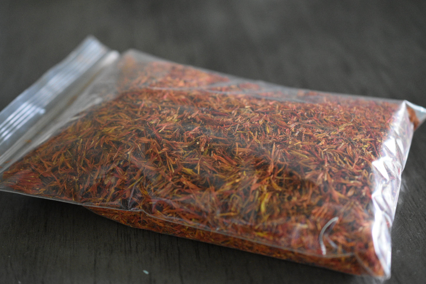 A plastic bag filled with safflower petals rests on a wooden surface to show the packaged product. The safflower petals are a vibrant red with hints of orange.