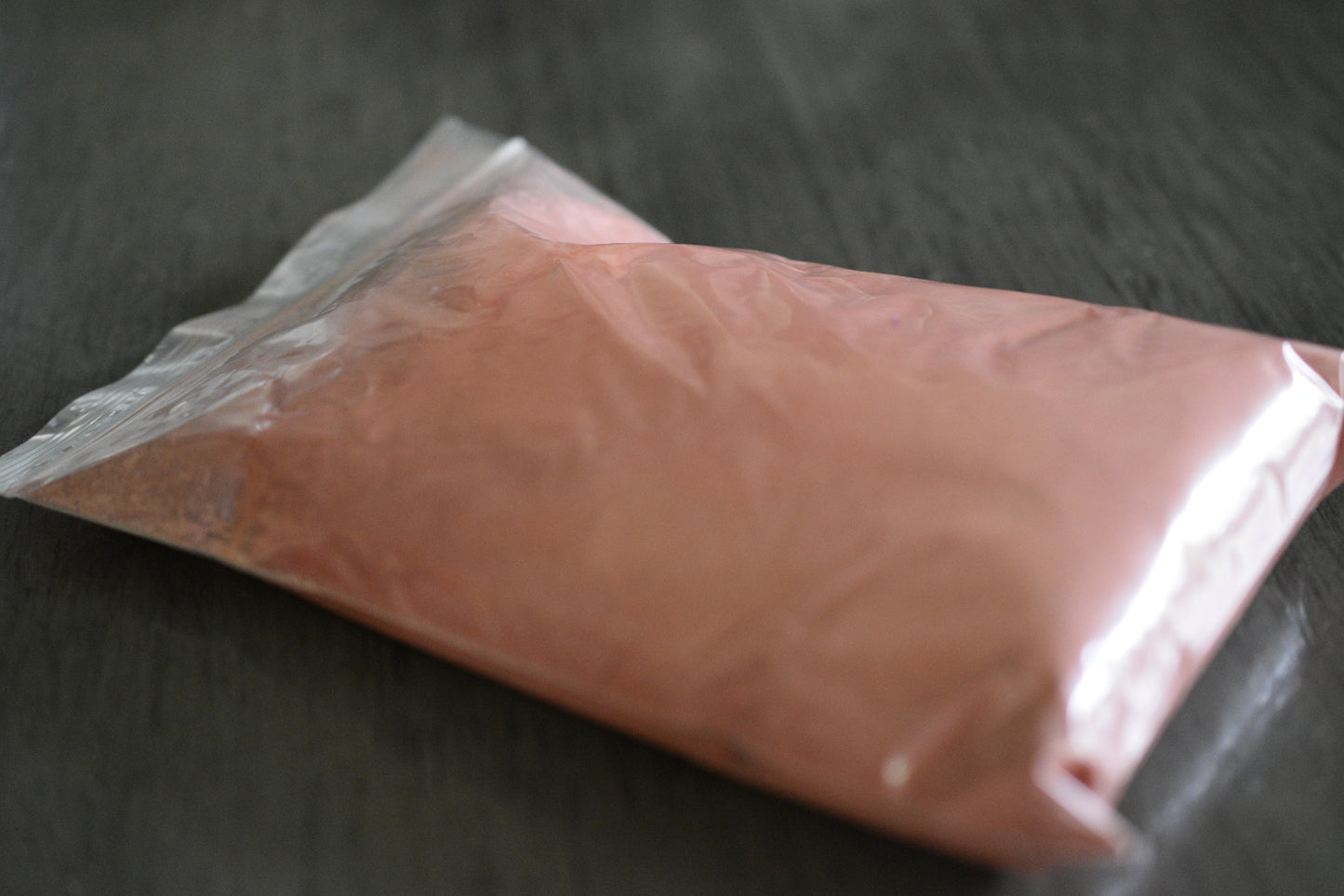 A plastic bag filled with rose clay rests on a wooden surface to show the packaged product. The clay is light pink in color.