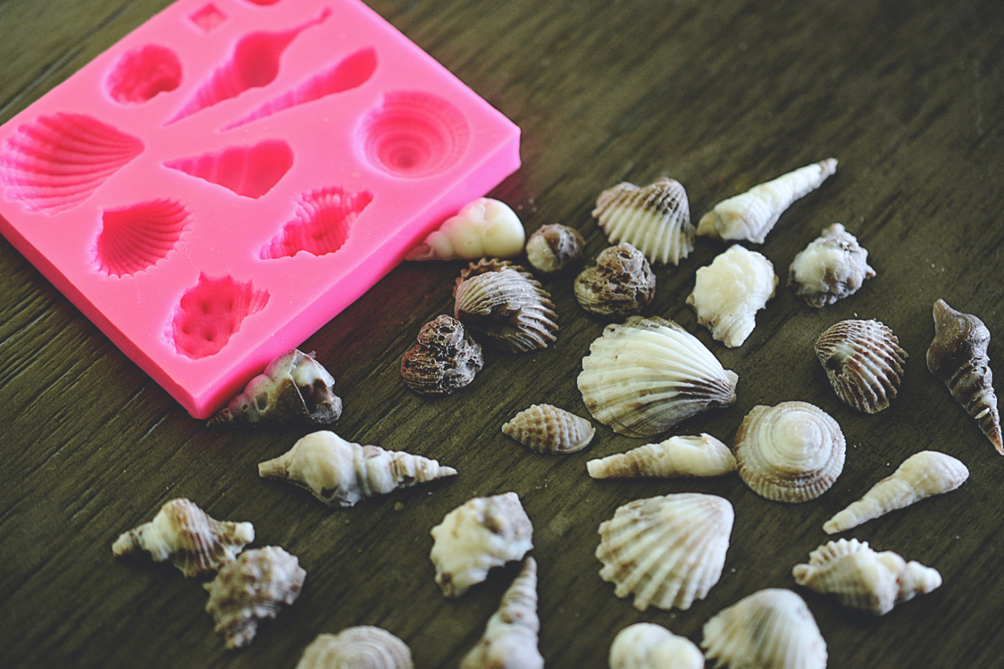 An assortment of seashells made from soap rests next to a square soap mold on a wooden surface. The seashells are colored a cream and brown color. The mold is pink.
