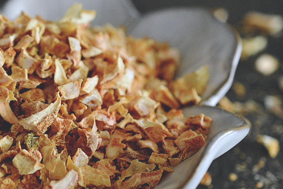 A close up view of an assortment of dried lemon peel rests in a white, flower shaped dish on a wooden surface. The lemon peel is a soft yellow and orange. There are small dried lemon peels scattered about in the background.