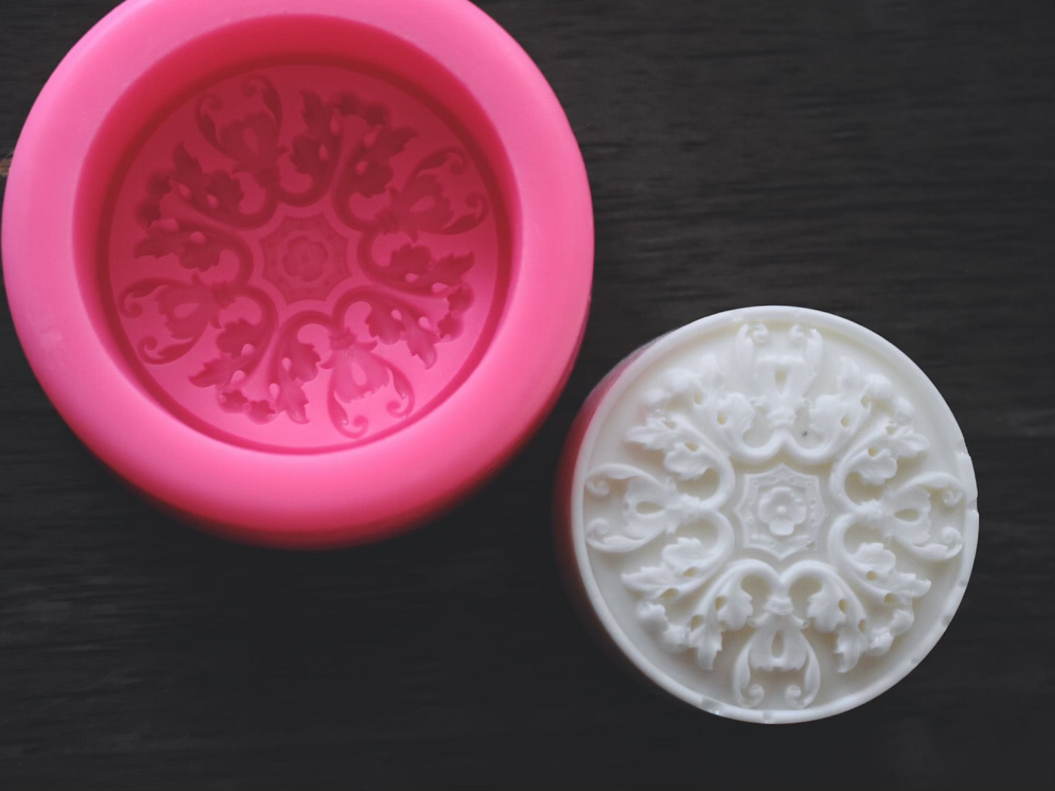 A round bar of soap with an elegant, filigree design rests next to round soap mold on a wooden surface. The mold is pink.