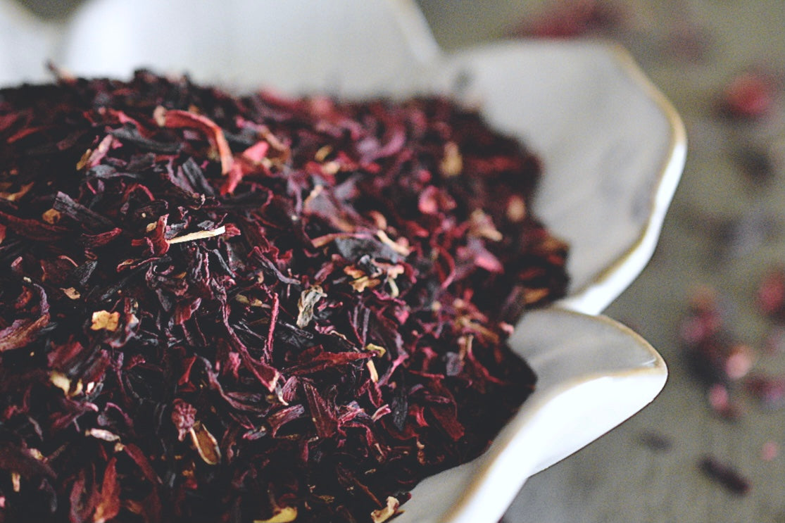 A close up view of dark red and black hibiscus flower petals rests in a white, flower shaped dish on a wooden surface. Hibiscus petals are scattered about in the background.