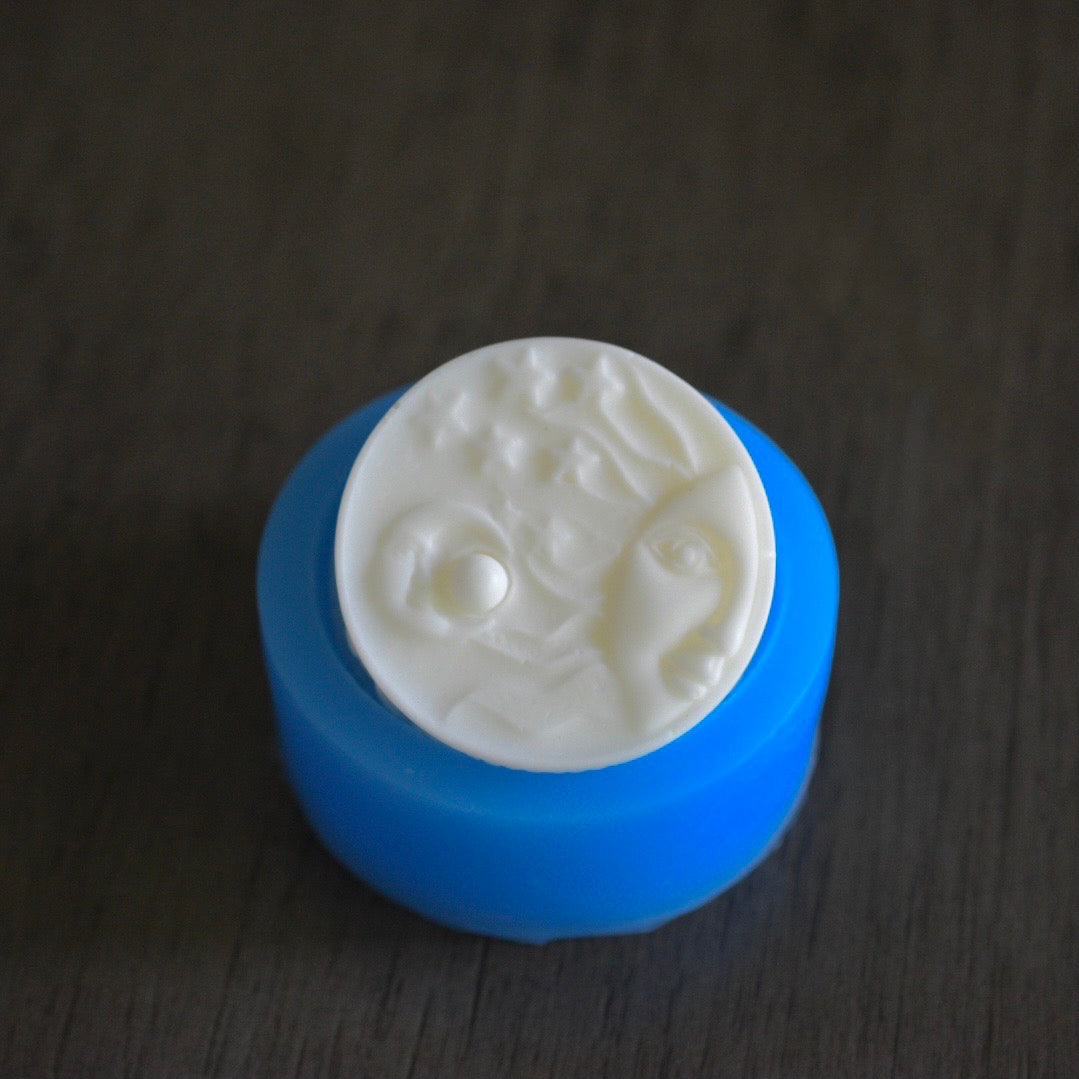 A round soap bar with a sun and moon design rests in a round soap mold on a wooden surface. The soap bar is white. The mold is blue.