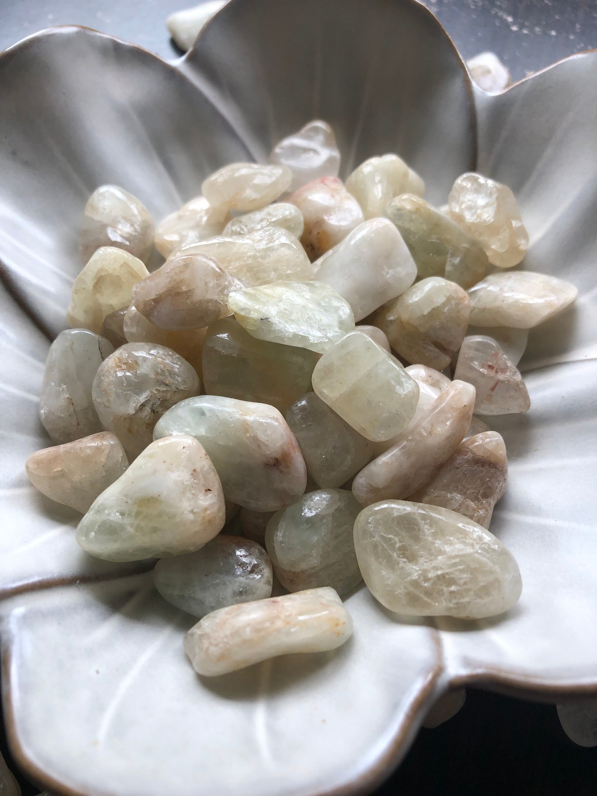 A white flower shaped bowl rests against a dark wood background. There are light brown and sandy colored tumbled stones in the bowl and scattered around the bowl for display.