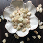 A white flower shaped bowl rests against a dark wood background. There are light brown and sandy colored tumbled stones in the bowl and scattered around the bowl for display.