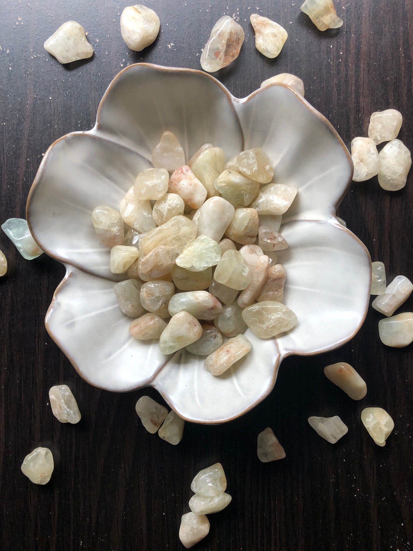 A white flower shaped bowl rests against a dark wood background. There are light brown and sandy colored tumbled stones in the bowl and scattered around the bowl for display.