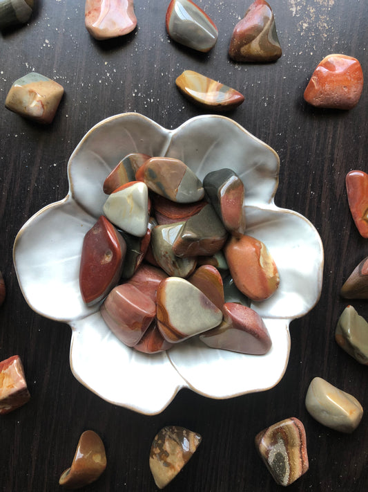 A white flower shaped bowl rests against a dark wood background. There are brown and red colored tumbled stones in the bowl and scattered around the bowl for display.