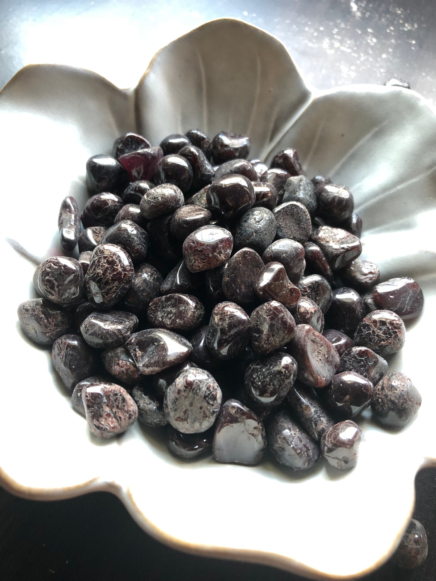 A white flower shaped bowl rests against a dark wood background. There are black and red flecked colored tumbled stones in the bowl and scattered around the bowl for display.