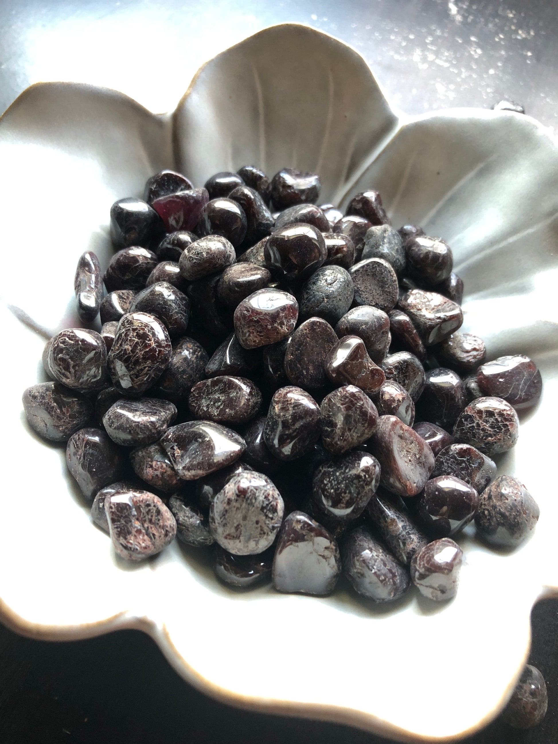 A white flower shaped bowl rests against a dark wood background. There are black and red flecked colored tumbled stones in the bowl and scattered around the bowl for display.