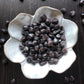 A white flower shaped bowl rests against a dark wood background. There are black and red flecked colored tumbled stones in the bowl and scattered around the bowl for display.