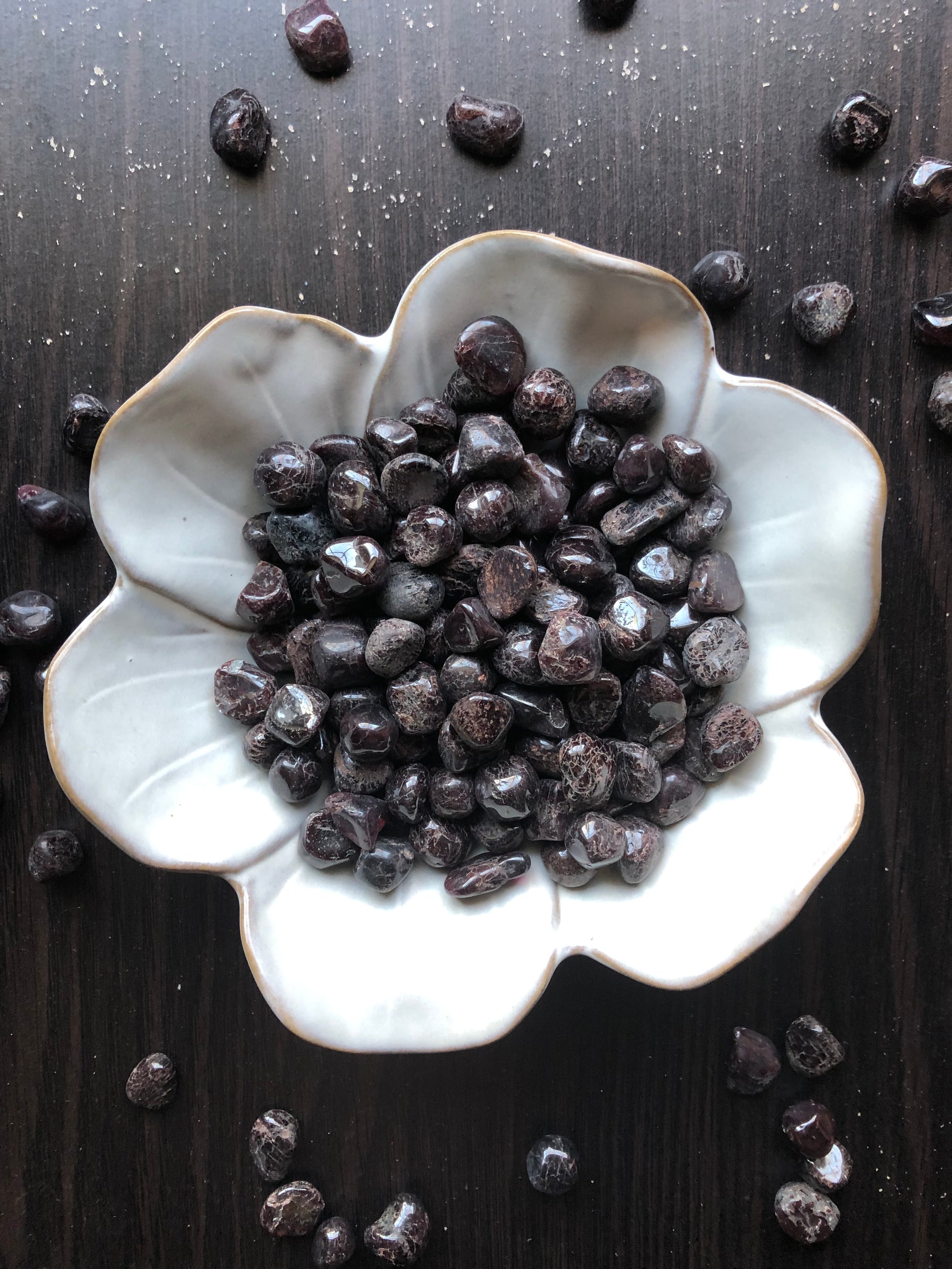 A white flower shaped bowl rests against a dark wood background. There are black and red flecked colored tumbled stones in the bowl and scattered around the bowl for display.