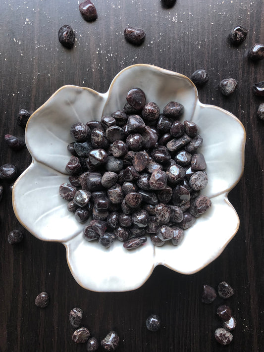 A white flower shaped bowl rests against a dark wood background. There are black and red flecked colored tumbled stones in the bowl and scattered around the bowl for display.