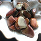 A white flower shaped bowl rests against a dark wood background. There are brown and red colored tumbled stones in the bowl and scattered around the bowl for display.