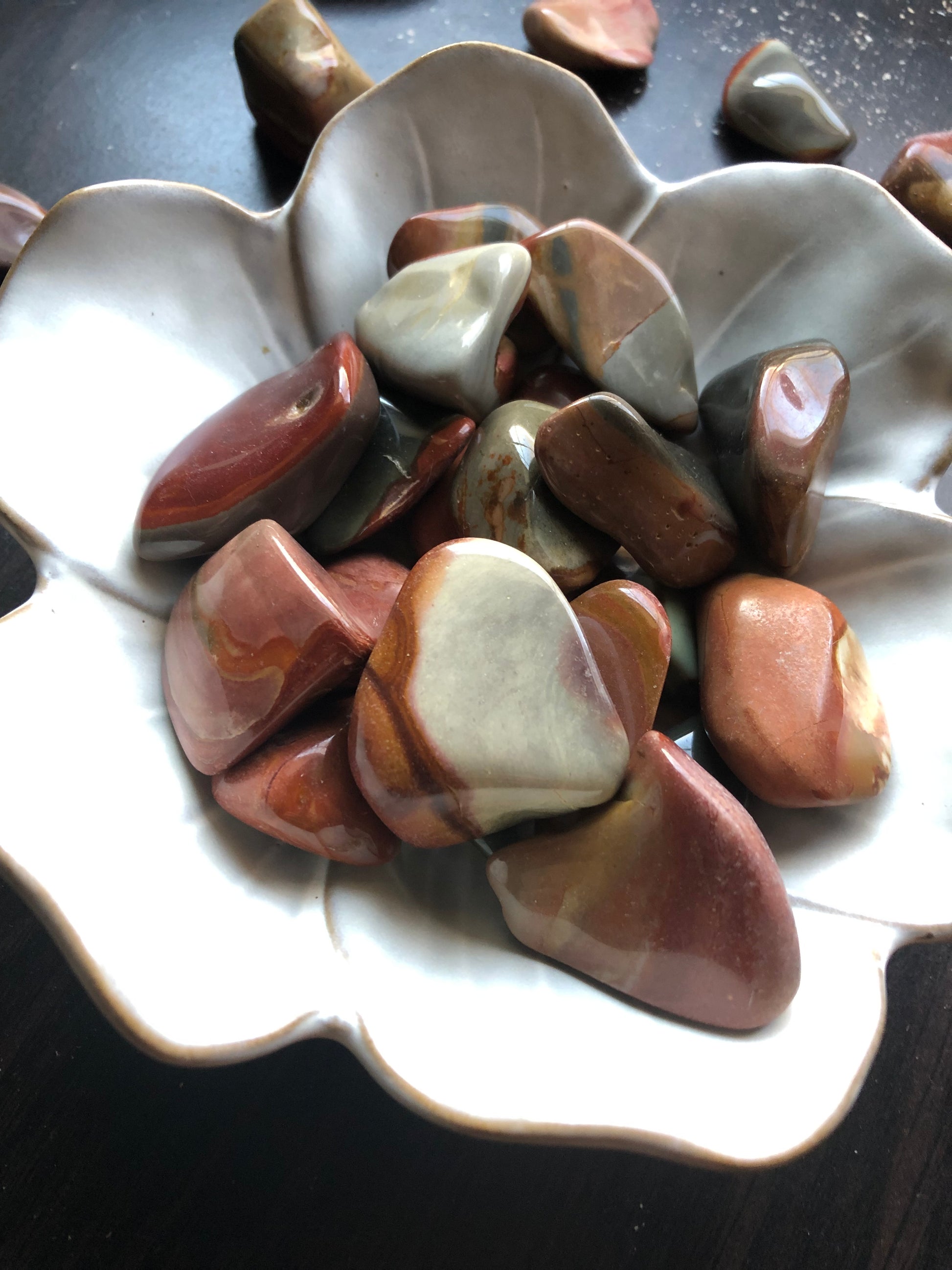 A white flower shaped bowl rests against a dark wood background. There are brown and red colored tumbled stones in the bowl and scattered around the bowl for display.