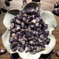 A downward picture of chevron banded amethyst stones sitting in a white, flower shaped bowl. There are chevron banded amethyst stones scattered around it. It sits on a dark wooden background.