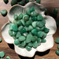 A downward picture of green aventurine stone sitting in a white, flower shaped bowl. There are scattered green aventurine stones around it. It sits on a dark wooden background.