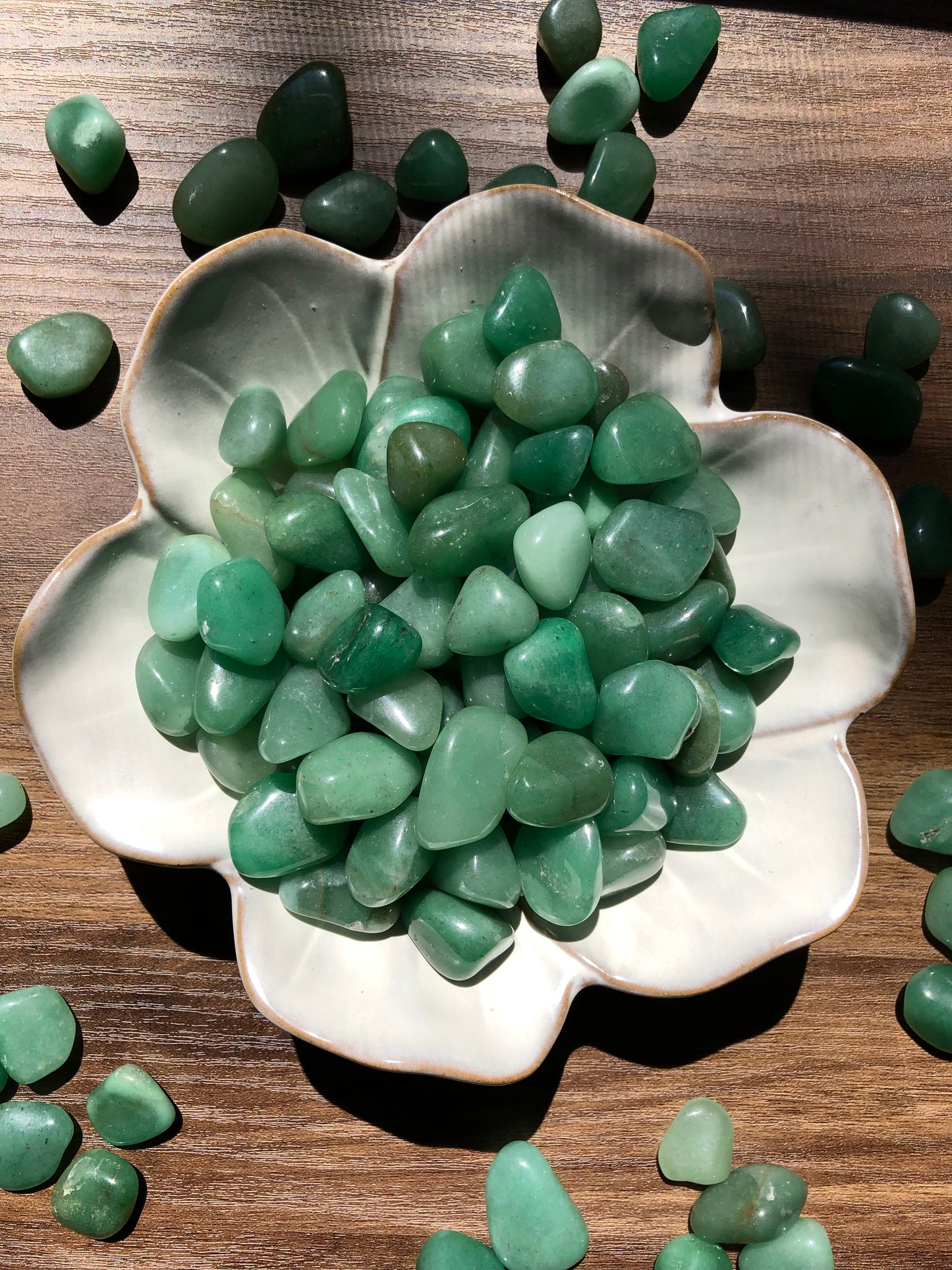 A downward picture of green aventurine stone sitting in a white, flower shaped bowl. There are scattered green aventurine stones around it. It sits on a dark wooden background.