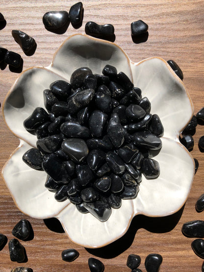 A downward picture of black onyx stones sitting in a white, flower shaped bowl. There are scattered black onyx stones around it. It sits on a dark wooden background.