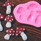 An assortment of mushrooms made from soap rests next to an oval soap mold on a wooden surface. The mushrooms are colored a white and red color. The mold has three slots in it. The mold is pink.