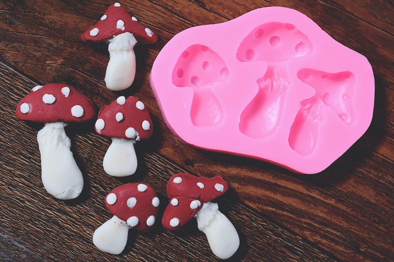 An assortment of mushrooms made from soap rests next to an oval soap mold on a wooden surface. The mushrooms are colored a white and red color. The mold has three slots in it. The mold is pink.