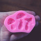 A hand holds a small pink mushroom mold against a wooden background. The mold has three slots in it. The mold is pink.