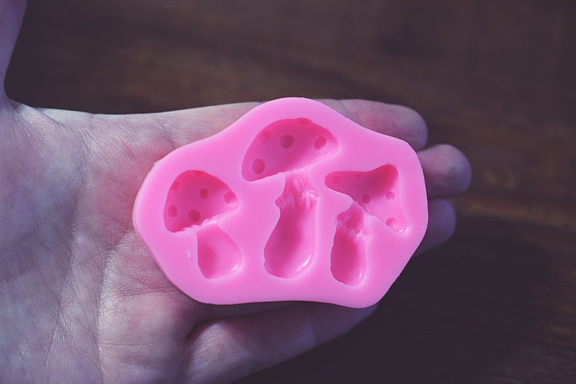 A hand holds a small pink mushroom mold against a wooden background. The mold has three slots in it. The mold is pink.