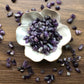 Tumbled amethyst stones of various shades of purple sit in a white bowl that is shaped like a flower with amethysts scattered around it. It sits on a dark wood background.