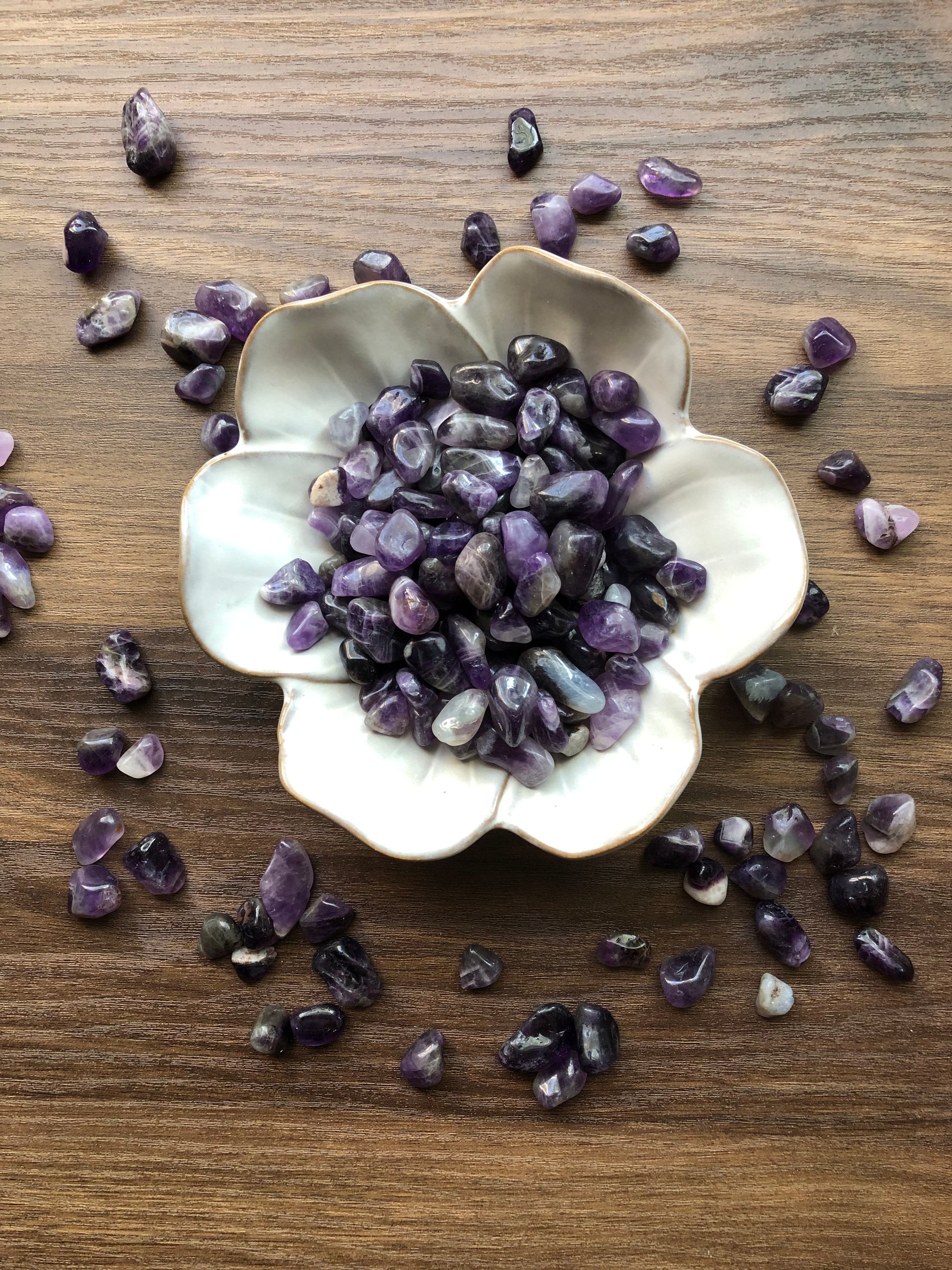 Tumbled amethyst stones of various shades of purple sit in a white bowl that is shaped like a flower with amethysts scattered around it. It sits on a dark wood background.