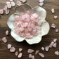 Tumbled rose quartz stones of various shades of pink sit in a white bowl that is shaped like a flower with rose quartz scattered around it. It sits on a dark wood background.