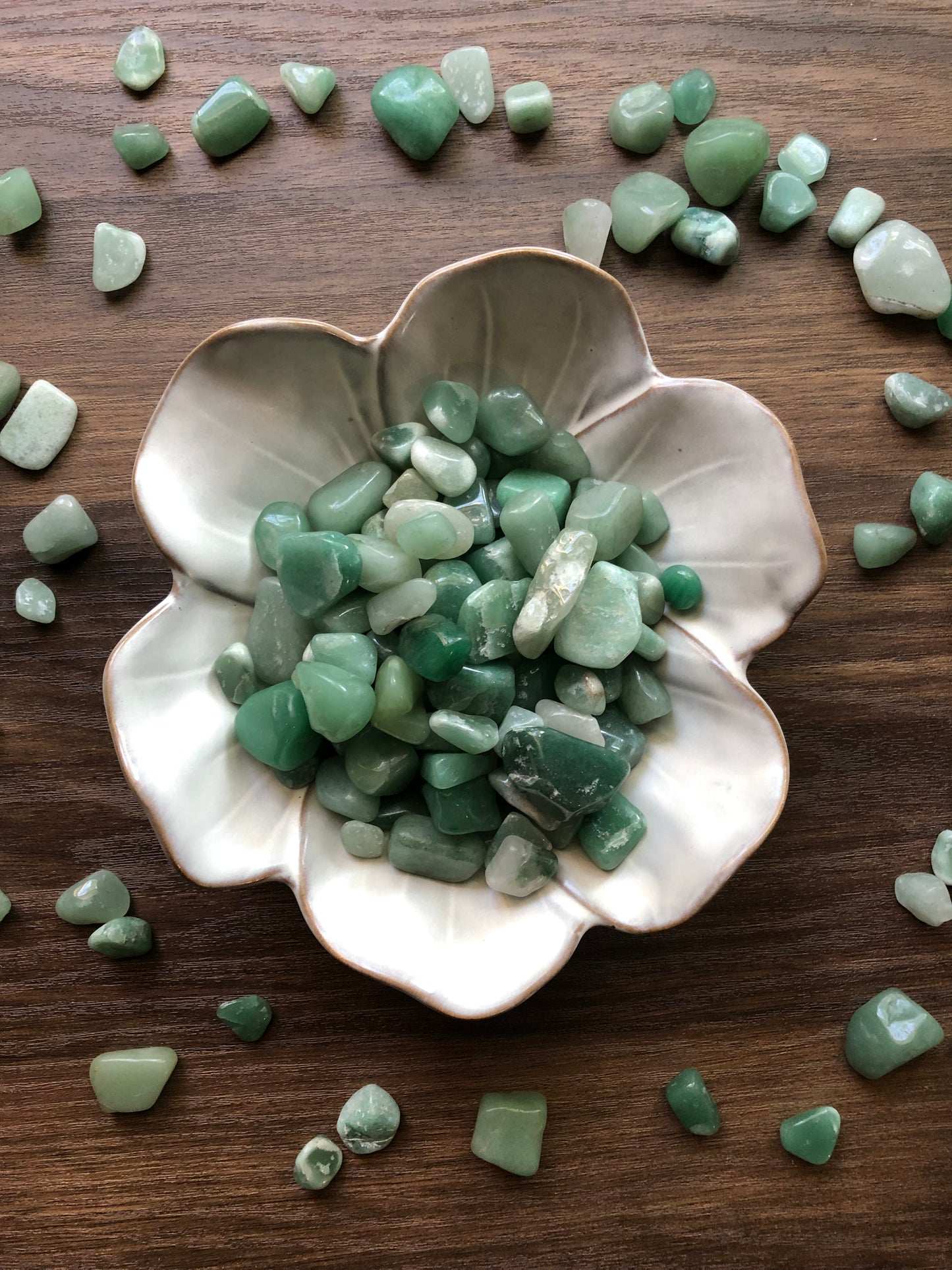 Tumbled aventurine stones of various shades of green sit in a white bowl that is shaped like a flower with aventurine scattered around it. It sits on a dark wood background.